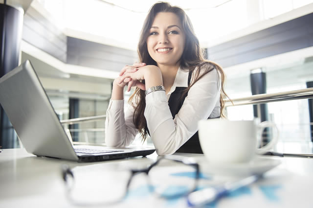 Imagem de uma mulher sentada em uma mesa, com um notebook e um óculos