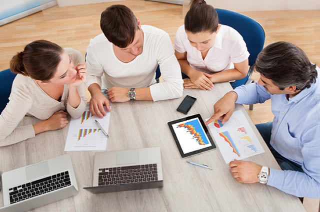 Mesa de reunião com 4 pessoas sentadas conversando e analisando sobre papéis contendo dados e gráficos